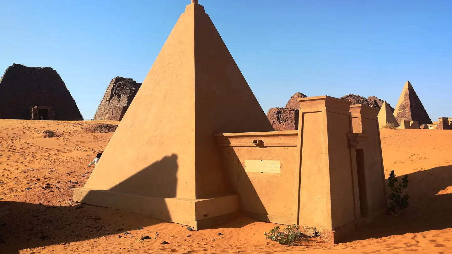 The pyramids of Meroe at Bagrawiyah. Steep sandstone pyramids and a chapel stand on red desert sand under a clear sky.